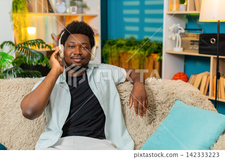 African American young man on home sofa enjoying music in headphones and nodding to rhythm indoors 142333223