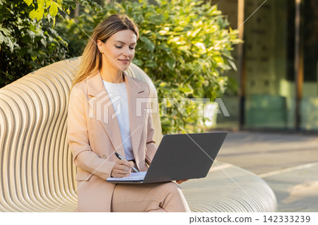 Young woman working on laptop in city street typing on bench near business center in windy weather Young woman working on laptop in city street typing on bench near business center in windy weather 142333239