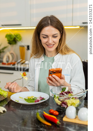Middle-aged woman enjoying salad while using smartphone in home kitchen for healthy diet lifestyle Middle-aged woman enjoying salad while using smartphone in home kitchen for healthy diet lifestyle 142333271