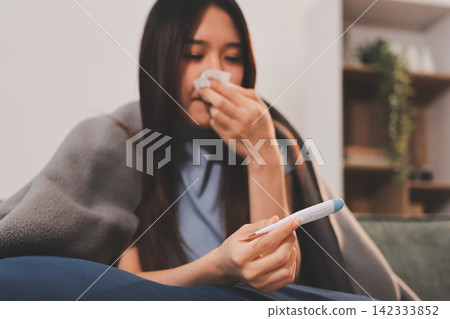 sick asian young woman sneezing into tissue paper while covered with a blanket and sitting on sofa in the living room sick asian young woman sneezing into tissue paper while covered with a blanket and sitting on sofa in the living room 142333852