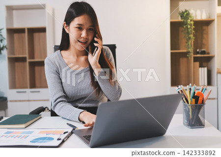 Young asian businesswoman is smiling and working on a laptop while sitting at her desk in a modern office Young asian businesswoman is smiling and working on a laptop while sitting at her desk in a modern office 142333892