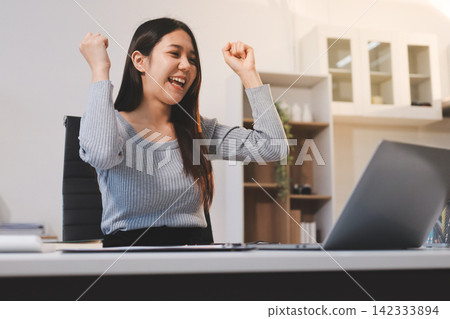 Young asian businesswoman is smiling and working on a laptop while sitting at her desk in a modern office Young asian businesswoman is smiling and working on a laptop while sitting at her desk in a modern office 142333894
