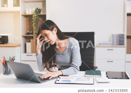 Young Asian woman sits at office desk rubbings her eyes and temples feeling pain. Stress. And exhaustion from long hours working on the computer Young Asian woman sits at office desk rubbings her eyes and temples feeling pain. Stress. And exhaustion from long hours working on the computer 142333895