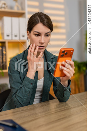 Young woman on home table seeing bad news on smartphone feeling worried shocked upset in home office Young woman on home table seeing bad news on smartphone feeling worried shocked upset in home office 142334235