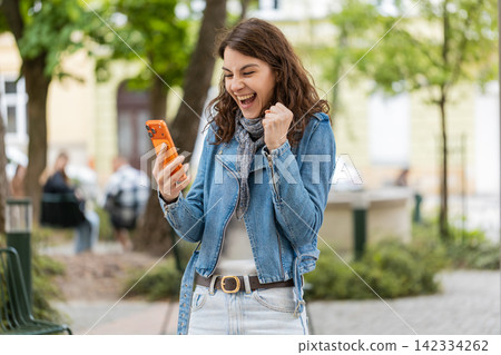 Young woman reading smartphone news walking in city street celebrating success with happy surprise Young woman reading smartphone news walking in city street celebrating success with happy surprise 142334262