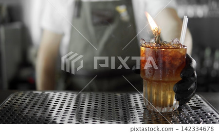 Bartender serving flaming craft cocktail with ice and garnish in a crystal glass on a bar counter Bartender serving flaming craft cocktail with ice and garnish in a crystal glass on a bar counter 142334678