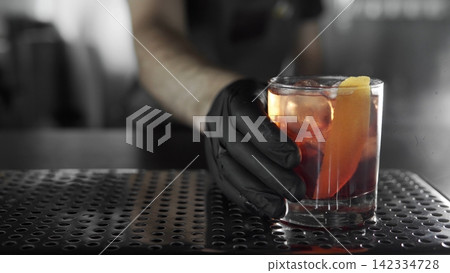 Bartender hand in black glove serving a negroni cocktail with orange peel and ice on a bar counter, offering professional service 142334728