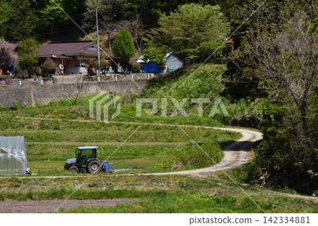 一輛農用拖拉機沿著農場道路穿過鄉村山區的田野和農田。 一輛農用拖拉機沿著農場道路穿過鄉村山區的田野和農田。 142334881