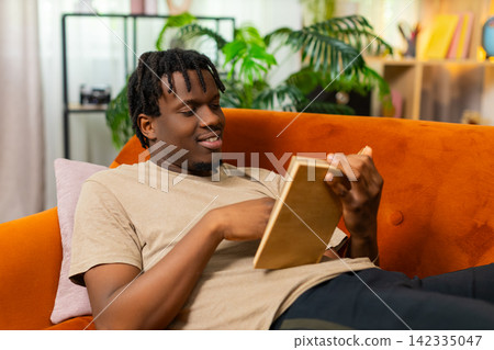 African American young man on home sofa reading book while lying on couch and turning page indoors African American young man on home sofa reading book while lying on couch and turning page indoors 142335047