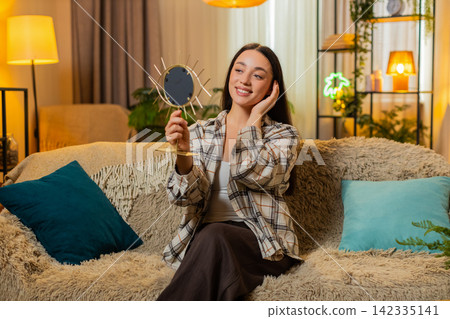 Young woman on home sofa checks look in small mirror fixes style and smiles with confidence at ease Young woman on home sofa checks look in small mirror fixes style and smiles with confidence at ease 142335141