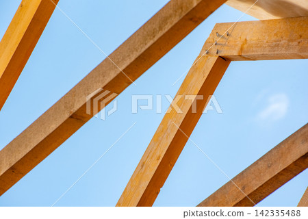 Wooden beams creating a skeletal structure of a roof truss, highlighting construction and architectural frameworks against the clear blue sky, symbolizing development and support Wooden beams creating a skeletal structure of a roof truss, highlighting construction and architectural frameworks against the clear blue sky, symbolizing development and support 142335488