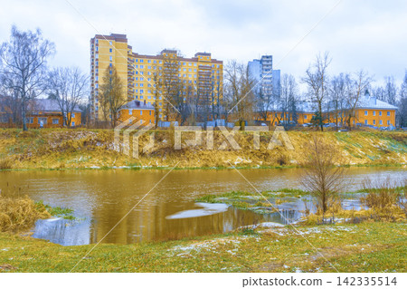 Riverside apartment buildings and yellow houses reflected in calm river under overcast sky Riverside apartment buildings and yellow houses reflected in calm river under overcast sky 142335514