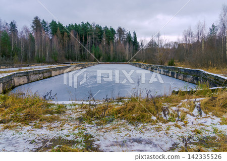 Abandoned concrete reservoir partially frozen surrounded by winter woodland under overcast sky Abandoned concrete reservoir partially frozen surrounded by winter woodland under overcast sky 142335526