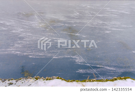Frozen lake surface with snow rim, mossy shoreline, pale winter light and open water patches Frozen lake surface with snow rim, mossy shoreline, pale winter light and open water patches 142335554