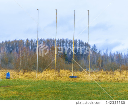 Four empty flagpoles standing on grassy field with autumn trees and pale overcast sky Four empty flagpoles standing on grassy field with autumn trees and pale overcast sky 142335580