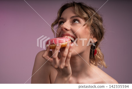 Young woman enjoying sweet pink iced donut, biting into dessert with happy expression, craving sugar and treat Young woman enjoying sweet pink iced donut, biting into dessert with happy expression, craving sugar and treat 142335594