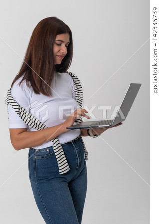 Young woman standing, holding a laptop and typing on it, representing concepts like online work, remote communication, digital learning, and modern technology use Young woman standing, holding a laptop and typing on it, representing concepts like online work, remote communication, digital learning, and modern technology use 142335739