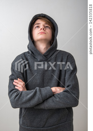 Young boy in a gray hoodie with crossed arms gazes upward in quiet contemplation, conveying dreaming, hope, concentration and future aspiration against a plain studio background 142335818