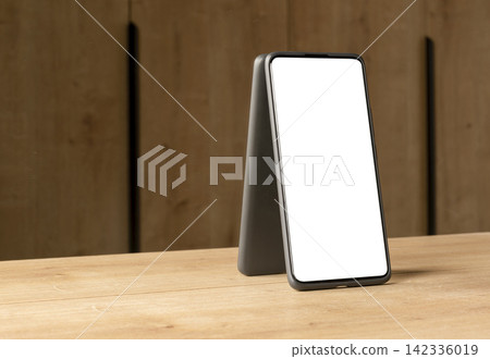 A smartphone with a blank white screen stands leaning against the corner of a dark wooden wall. The A smartphone with a blank white screen stands leaning against the corner of a dark wooden wall. The 142336019