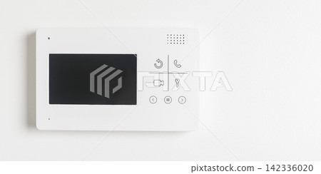 A wall-mounted video intercom panel with a small screen and call buttons sits against a white A wall-mounted video intercom panel with a small screen and call buttons sits against a white 142336020