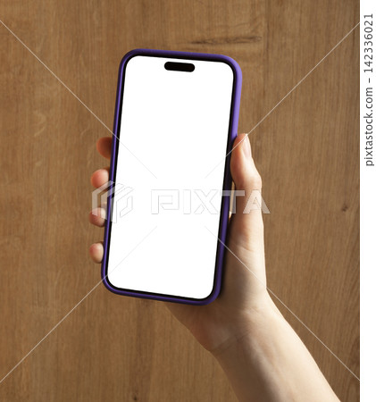 A woman's hand holds a smartphone with a blank white screen against a wooden wall. The mobile mockup A woman's hand holds a smartphone with a blank white screen against a wooden wall. The mobile mockup 142336021