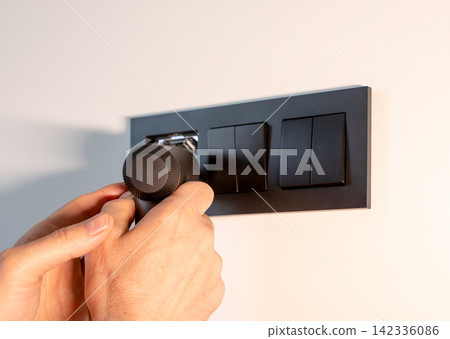 Hand removes or installs the inner mechanism of a black electrical socket on a white wall. The 142336086