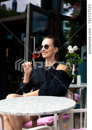 Stylish woman in sunglasses enjoys rose champagne at outdoor cafe, smiling while holding elegant glass near flowers. Relaxed summer autumn portrait leisure, luxury, fashion, travel, urban lifestyle Stylish woman in sunglasses enjoys rose champagne at outdoor cafe, smiling while holding elegant glass near flowers. Relaxed summer autumn portrait leisure, luxury, fashion, travel, urban lifestyle 142337523