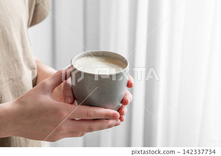 Human hands holding a cup of cappuccino coffee on a window background with morning shadow 142337734