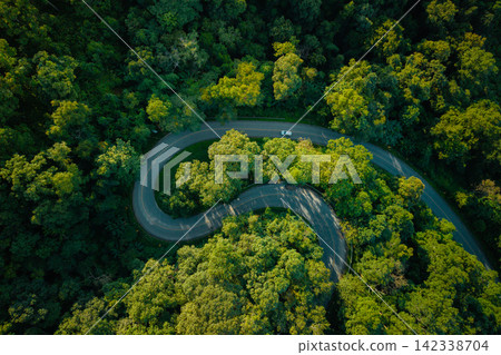 Aerial view of a winding road through a lush green forest with a driving car. Aerial view of a winding road through a lush green forest with a driving car. 142338704