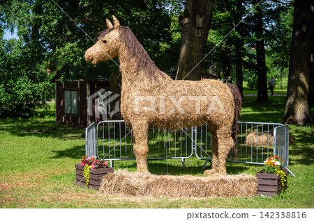 A large horse made of straw sits in a green park with trees on a sunny day with flowers around. A large horse made of straw sits in a green park with trees on a sunny day with flowers around. 142338816