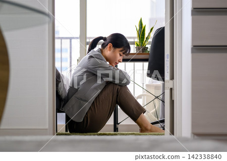一位年輕女子沮喪地坐在房間裡，抱著膝蓋。 142338840