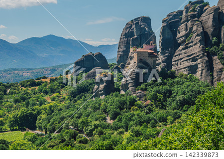 Panoramic view of Meteora's towering rock formations surrounded by dense green vegetation and distant mountain ranges, featuring ancient monasteries built on the cliffs in a serene landscape Panoramic view of Meteora's towering rock formations surrounded by dense green vegetation and distant mountain ranges, featuring ancient monasteries built on the cliffs in a serene landscape 142339873