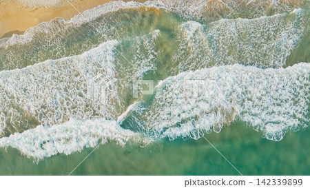 Aerial perspective of ocean waves breaking on a sandy beach, with clear turquoise water and white foam visible, creating a vibrant contrast against the shoreline under bright sunlight Aerial perspective of ocean waves breaking on a sandy beach, with clear turquoise water and white foam visible, creating a vibrant contrast against the shoreline under bright sunlight 142339899