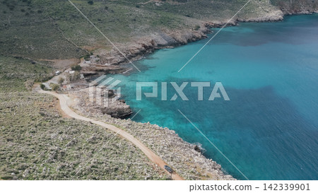 Aerial view of a winding coastal road approaching Ombros Gialos Bay in Crete, featuring clear turquoise waters, rocky cliffs, and verdant hills, creating a picturesque natural landscape Aerial view of a winding coastal road approaching Ombros Gialos Bay in Crete, featuring clear turquoise waters, rocky cliffs, and verdant hills, creating a picturesque natural landscape 142339901