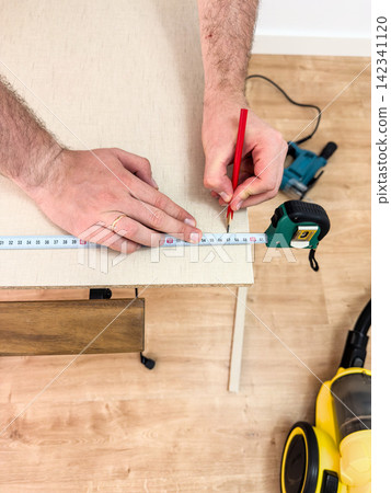 Man marks a cutting length on a chipboard panel beside a jigsaw Man marks a cutting length on a chipboard panel beside a jigsaw 142341120
