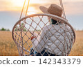 Woman reading printed book on hanging swing overlooking a wheat field at sunset 142343379