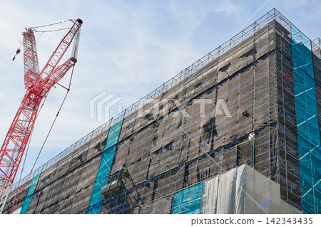 The exterior of a high-rise building under construction and a large crane reaching towards the blue sky. The exterior of a high-rise building under construction and a large crane reaching towards the blue sky. 142343435