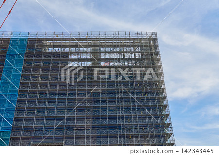 在湛藍的天空下,一座正在興建中的高層建築外牆被鷹架和防護布覆蓋。 在湛藍的天空下,一座正在興建中的高層建築外牆被鷹架和防護布覆蓋。 142343445