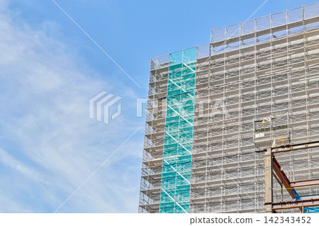 The exterior of a high-rise building under construction, covered in scaffolding and protective sheeting, under a clear blue sky. The exterior of a high-rise building under construction, covered in scaffolding and protective sheeting, under a clear blue sky. 142343452