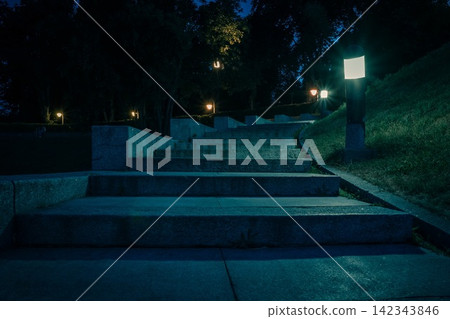 The tiled stairs in the night green park with lanterns in summer. Benches in the park during the summer season at night. Illumination of a park road with lanterns. Park of Eternal Glory. Ukraine The tiled stairs in the night green park with lanterns in summer. Benches in the park during the summer season at night. Illumination of a park road with lanterns. Park of Eternal Glory. Ukraine 142343846