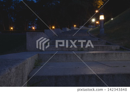 The tiled stairs in the night green park with lanterns in summer. Benches in the park during the summer season at night. Illumination of a park road with lanterns. Park of Eternal Glory. Ukraine The tiled stairs in the night green park with lanterns in summer. Benches in the park during the summer season at night. Illumination of a park road with lanterns. Park of Eternal Glory. Ukraine 142343848