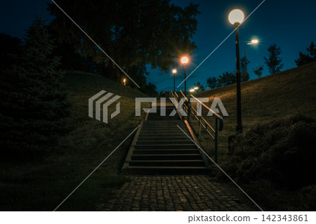 The tiled stairs in the night green park with lanterns in summer. Benches in the park during the summer season at night. Illumination of a park road with lanterns. Park of Eternal Glory. Ukraine The tiled stairs in the night green park with lanterns in summer. Benches in the park during the summer season at night. Illumination of a park road with lanterns. Park of Eternal Glory. Ukraine 142343861