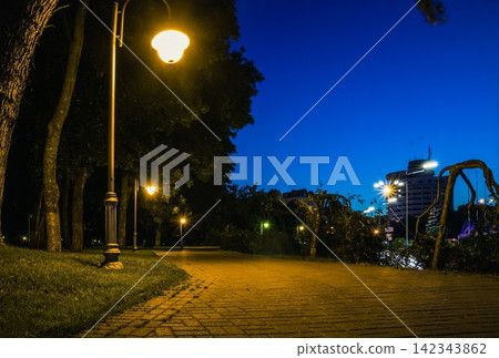 The tiled road in the night green park with lanterns in summer. Benches in the park during the summer season at night. Illumination of a park road with lanterns. Park of Eternal Glory. Ukraine The tiled road in the night green park with lanterns in summer. Benches in the park during the summer season at night. Illumination of a park road with lanterns. Park of Eternal Glory. Ukraine 142343862