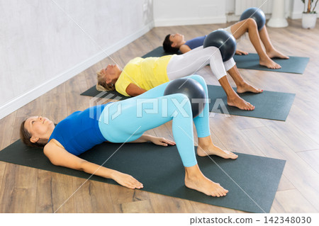 Middle-aged woman lying on fitness rug with softball between legs in training room Middle-aged woman lying on fitness rug with softball between legs in training room 142348030