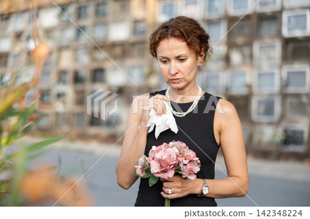 Sad woman in black dress with bouquet of flowers and handkerchief in hands, stands in cemetery Sad woman in black dress with bouquet of flowers and handkerchief in hands, stands in cemetery 142348224
