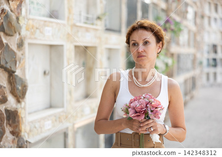Sad woman in white croptop with bouquet of flowers and handkerchief in hands, stands in cemetery Sad woman in white croptop with bouquet of flowers and handkerchief in hands, stands in cemetery 142348312