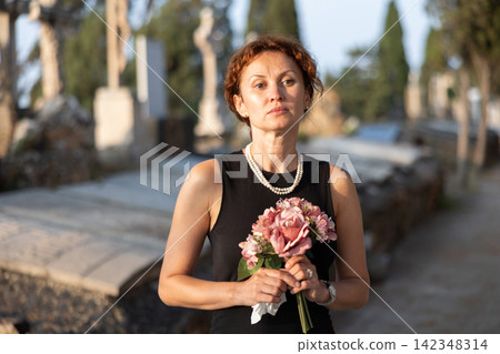 Woman in black dress with bouquet of flowers came to cemetery to honor the memory of deceased loved one Woman in black dress with bouquet of flowers came to cemetery to honor the memory of deceased loved one 142348314