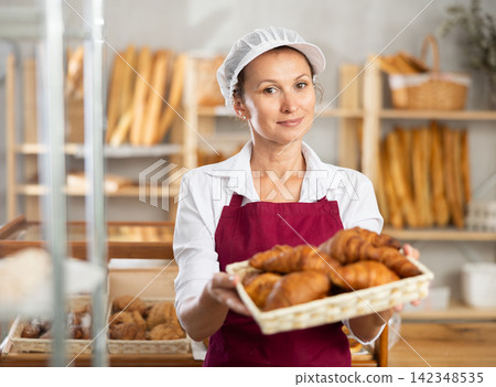 Female baker in white uniform stands behind counter in small private bakery offering hot croissants Female baker in white uniform stands behind counter in small private bakery offering hot croissants 142348535