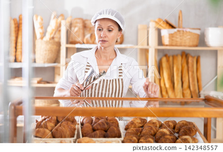 Selling fresh croissants in bakery - female baker places hot croissants on display Selling fresh croissants in bakery - female baker places hot croissants on display 142348557
