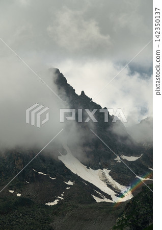 Dramatic mountain peak obscured by clouds with snow and rainbow Dramatic mountain peak obscured by clouds with snow and rainbow 142350137